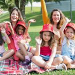 Two women and four girls enjoy a sunny picnic in Portugal, waving happily in the park.